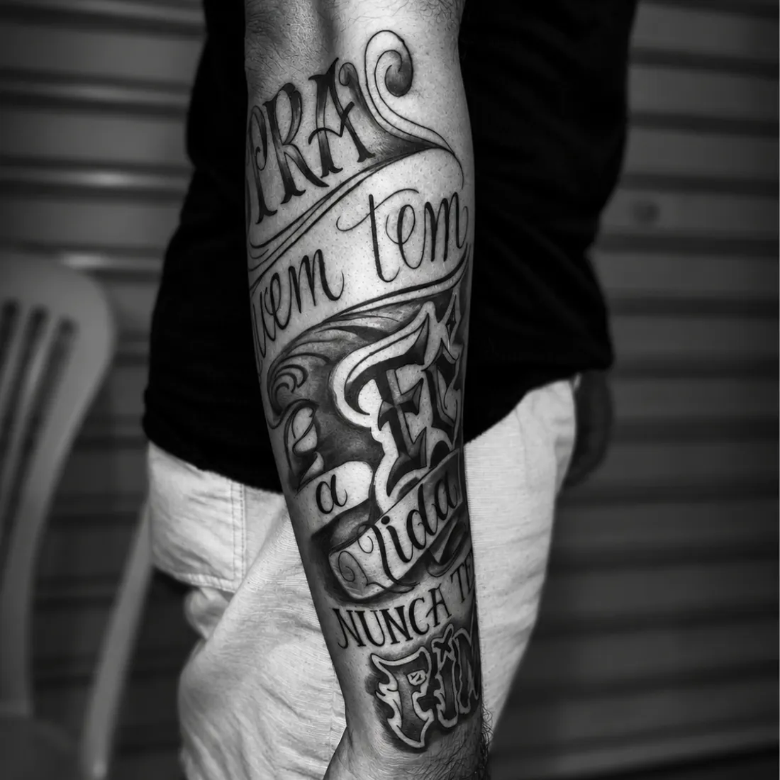 Tatuagem autoral de lettering em faixas envolvendo o antebraço, em preto e cinza