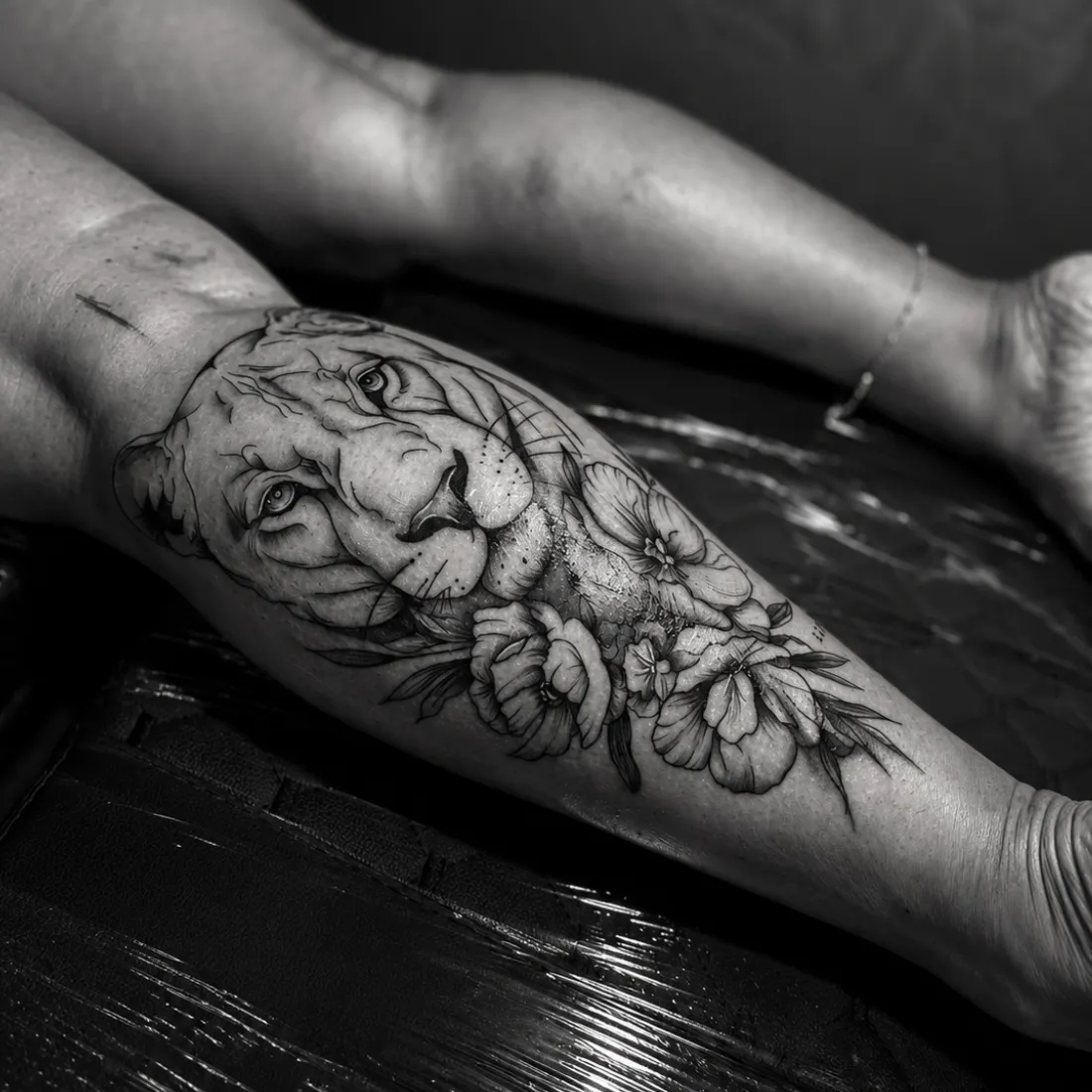 Tatuagem de traço fino com rosto de leoa e flores na perna, em preto e branco